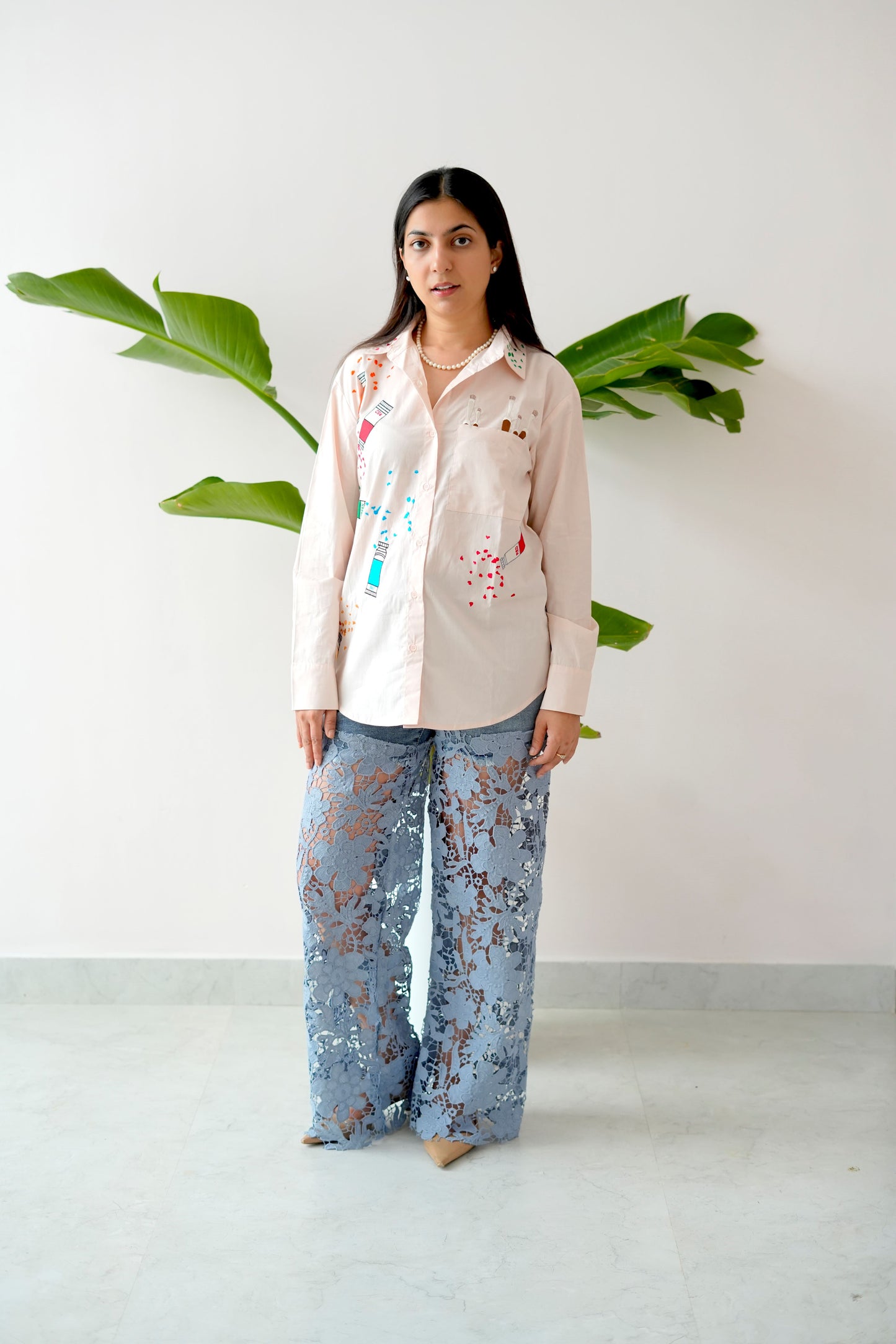 Woman wearing a light-colored blouse with floral patterns and light blue floral pants, standing against a white wall with green leaves.
