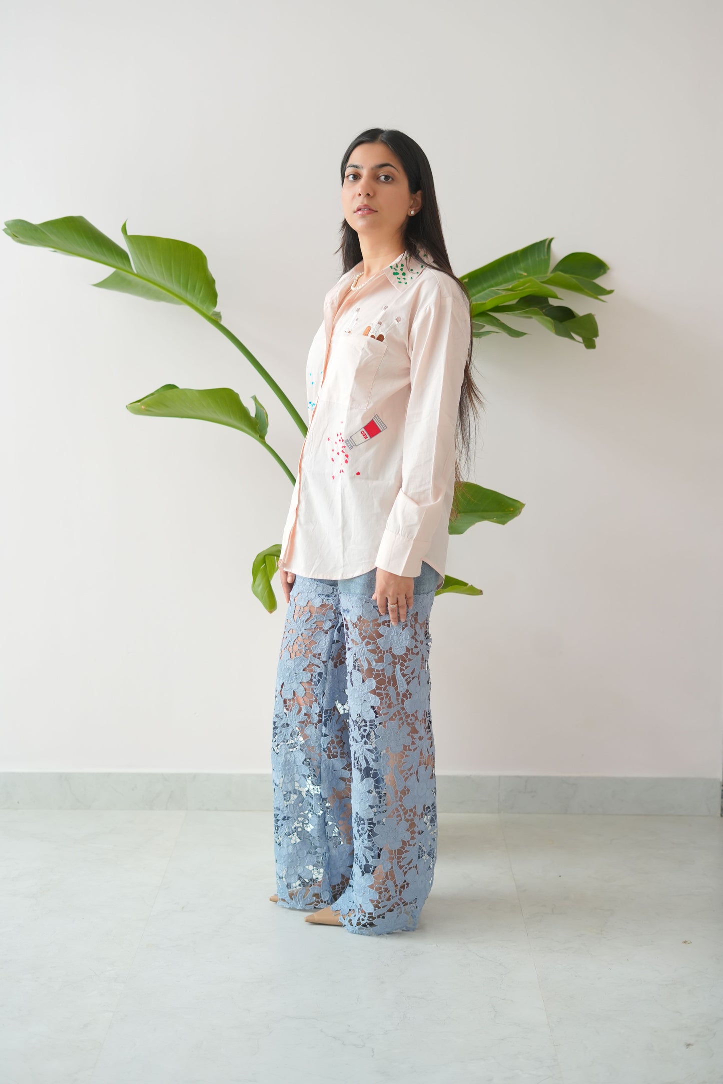 Woman wearing a white blouse with floral embroidery and blue patterned pants standing against a white wall.