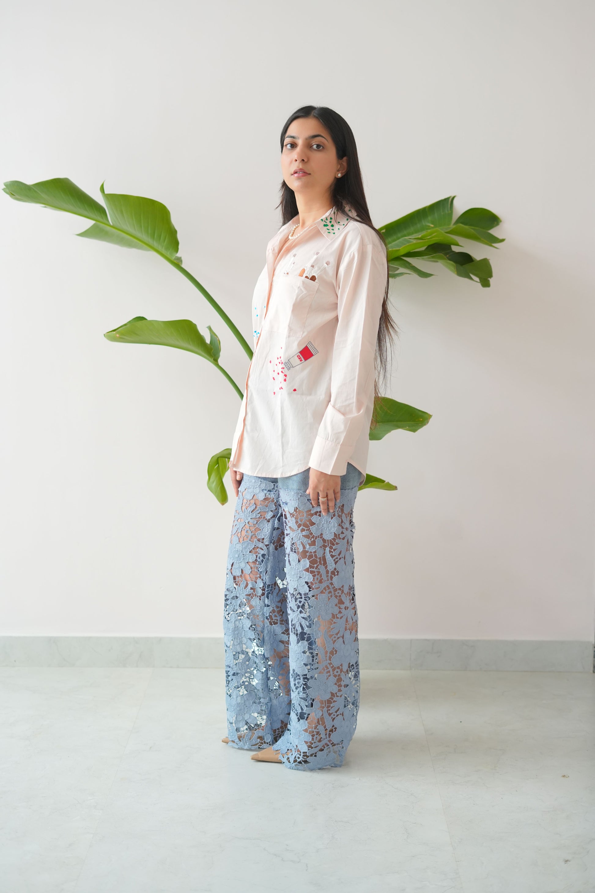 Woman wearing a white blouse with floral embroidery and blue patterned pants standing against a white wall.
