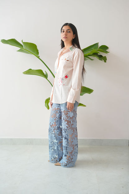 Woman wearing a white blouse with floral embroidery and blue patterned pants standing against a white wall.
