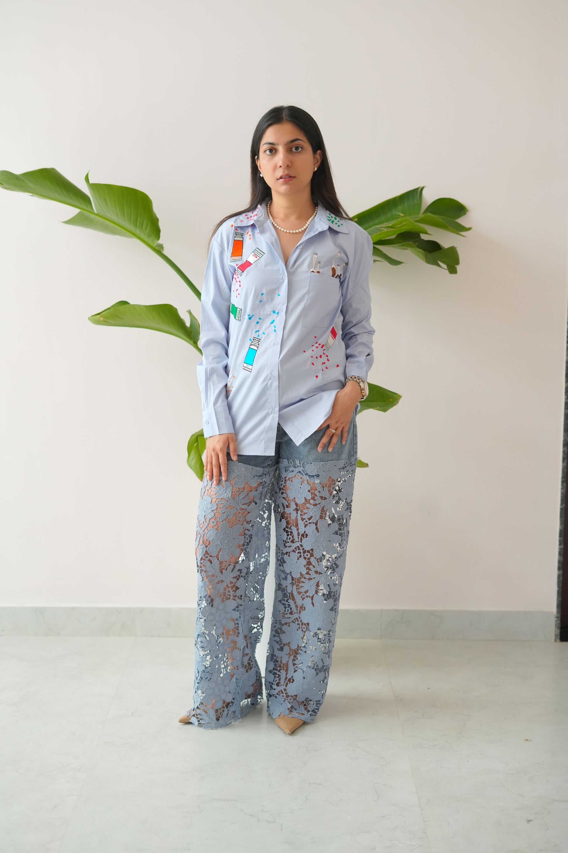 Person wearing a light gray hoodie and floral pants standing next to a plant indoors.
