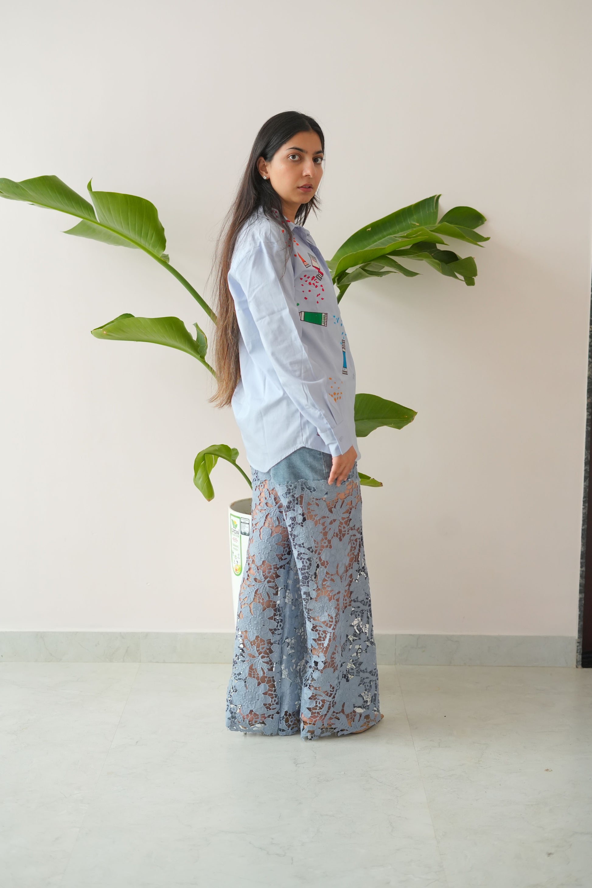 Person wearing a light-colored outfit with floral pants standing next to a plant indoors.