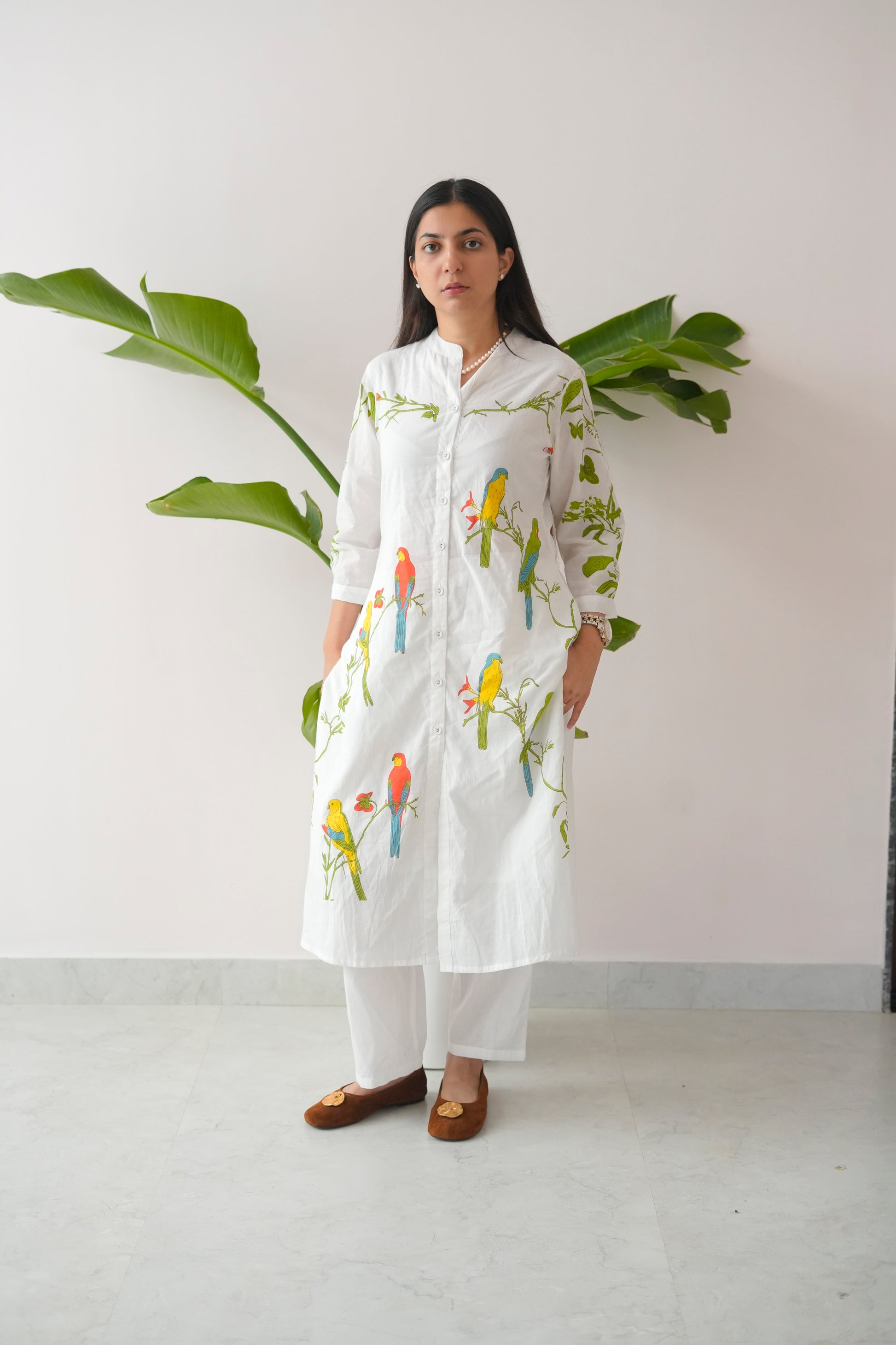 Woman wearing a white kurta with bird prints standing against a white wall with a plant.