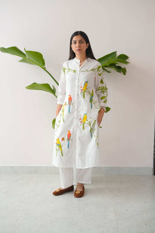 Woman wearing a white kurta with bird prints standing against a white wall with a plant.