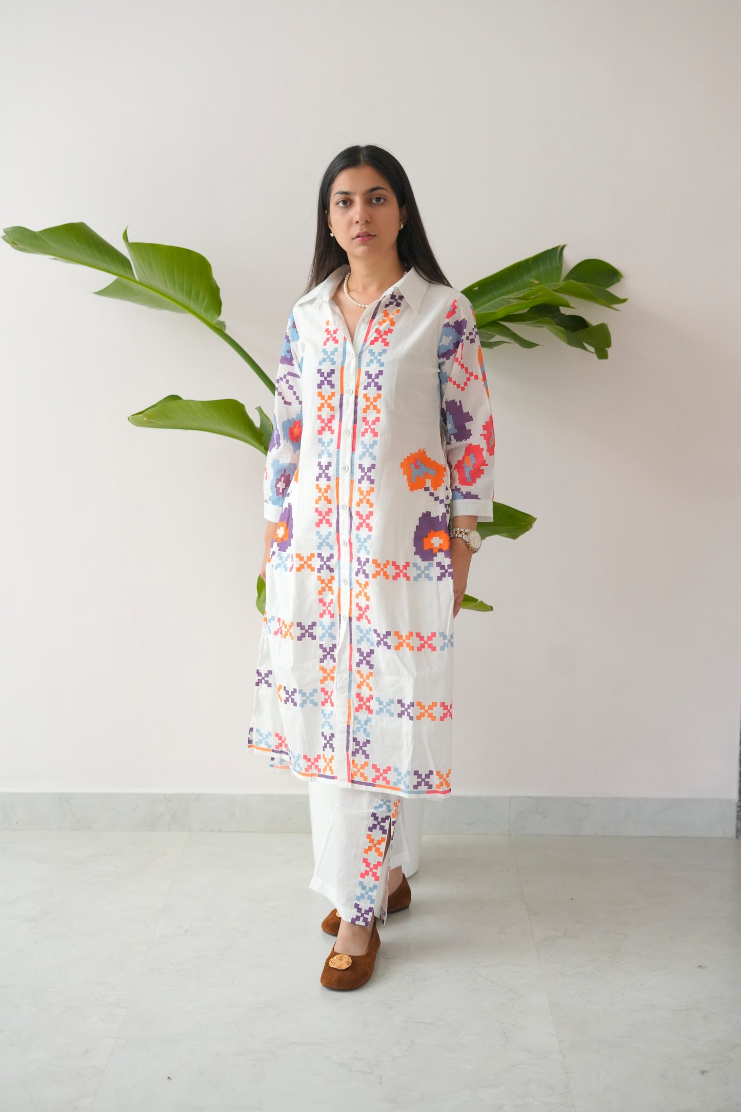 Woman wearing a colorful embroidered kurta with white pants against a plain background