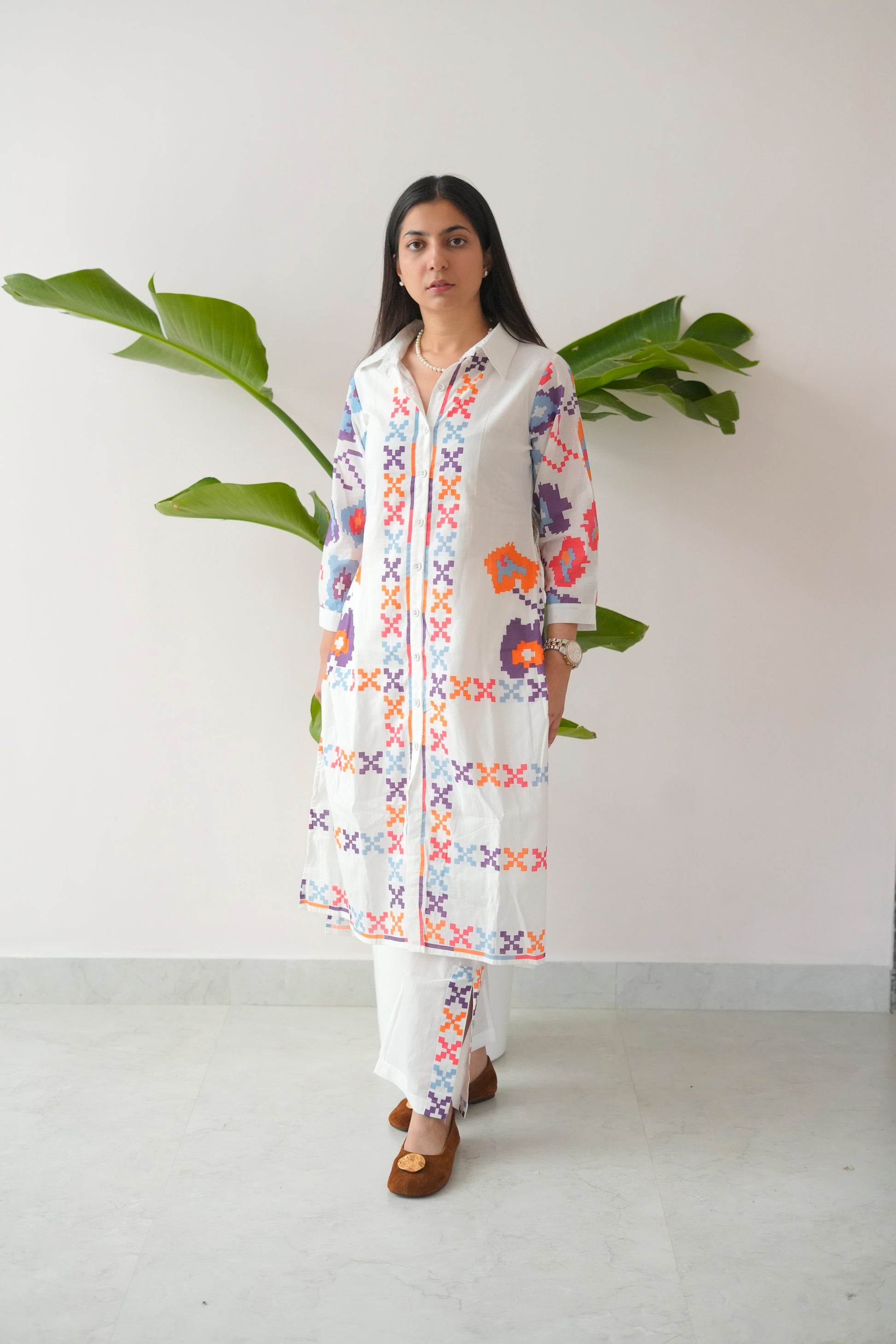 Woman wearing a colorful embroidered kurta with white pants against a plain background