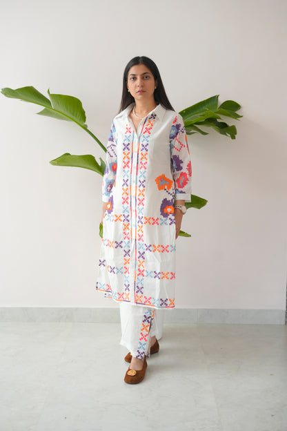 Woman wearing a colorful embroidered kurta with white pants against a plain background