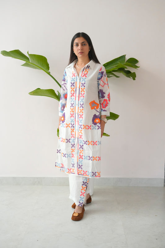 Woman wearing a colorful embroidered kurta with white pants against a plain background