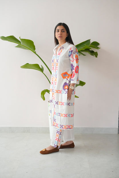 Woman wearing a long, white embroidered dress with colorful patterns against a plain background.