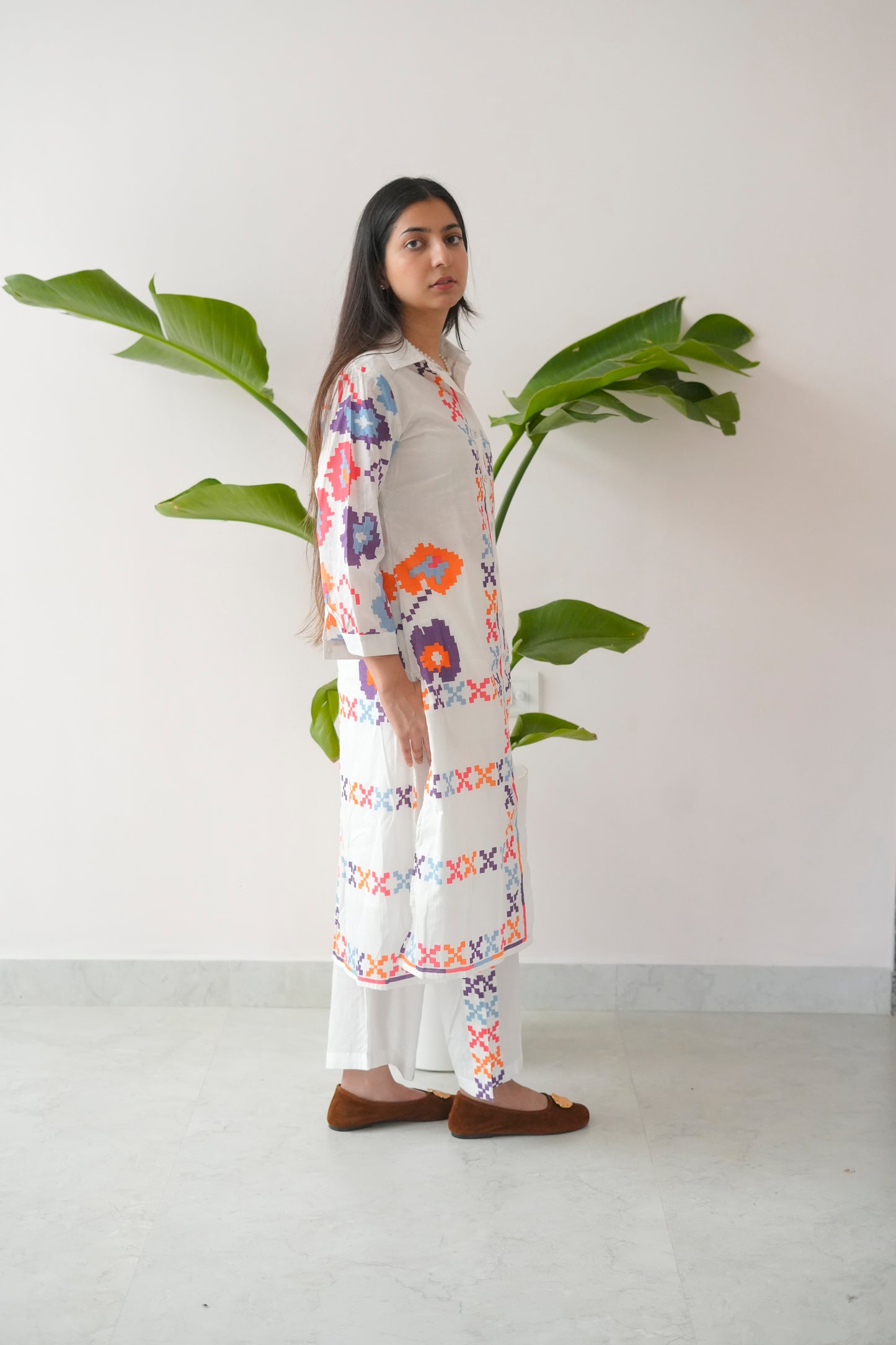 Woman wearing a floral dress standing next to a plant against a white wall.