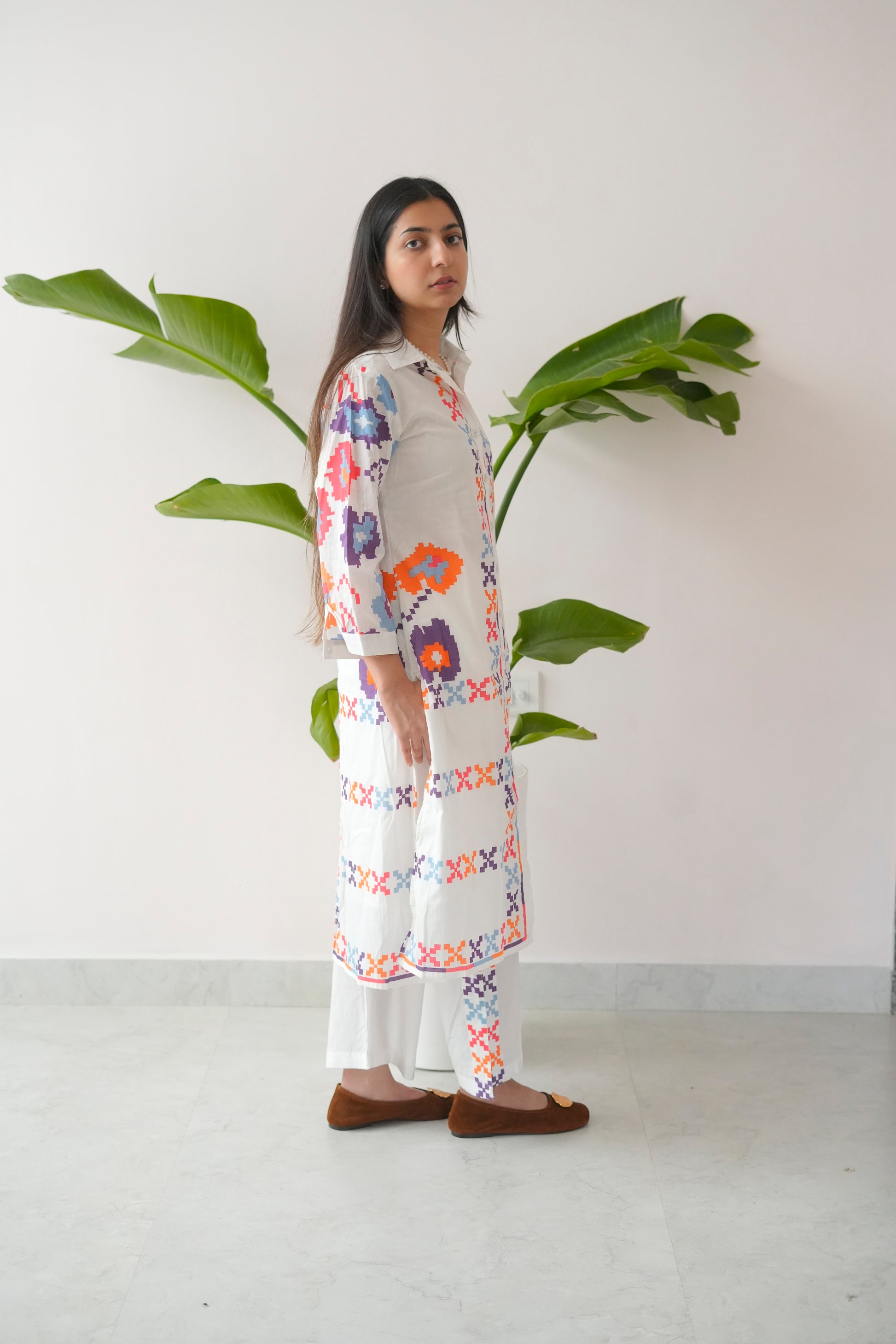Woman wearing a floral dress standing next to a plant against a white wall.