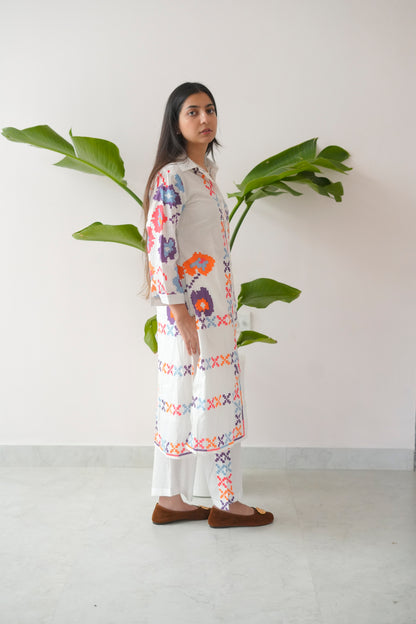 Woman wearing a floral dress standing next to a plant against a white wall.