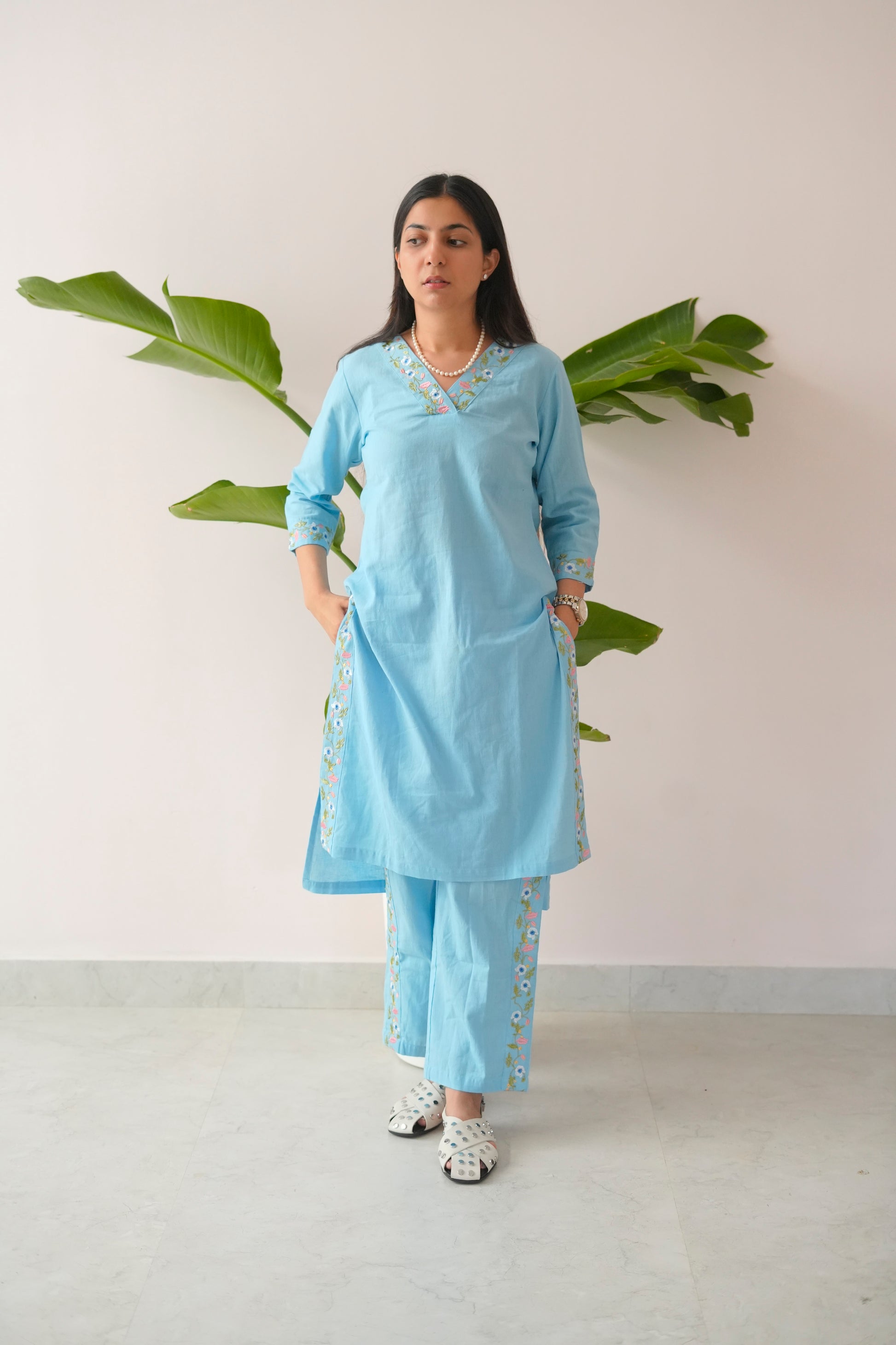 Woman wearing a light blue outfit with white patterns standing against a plain background with green leaves.