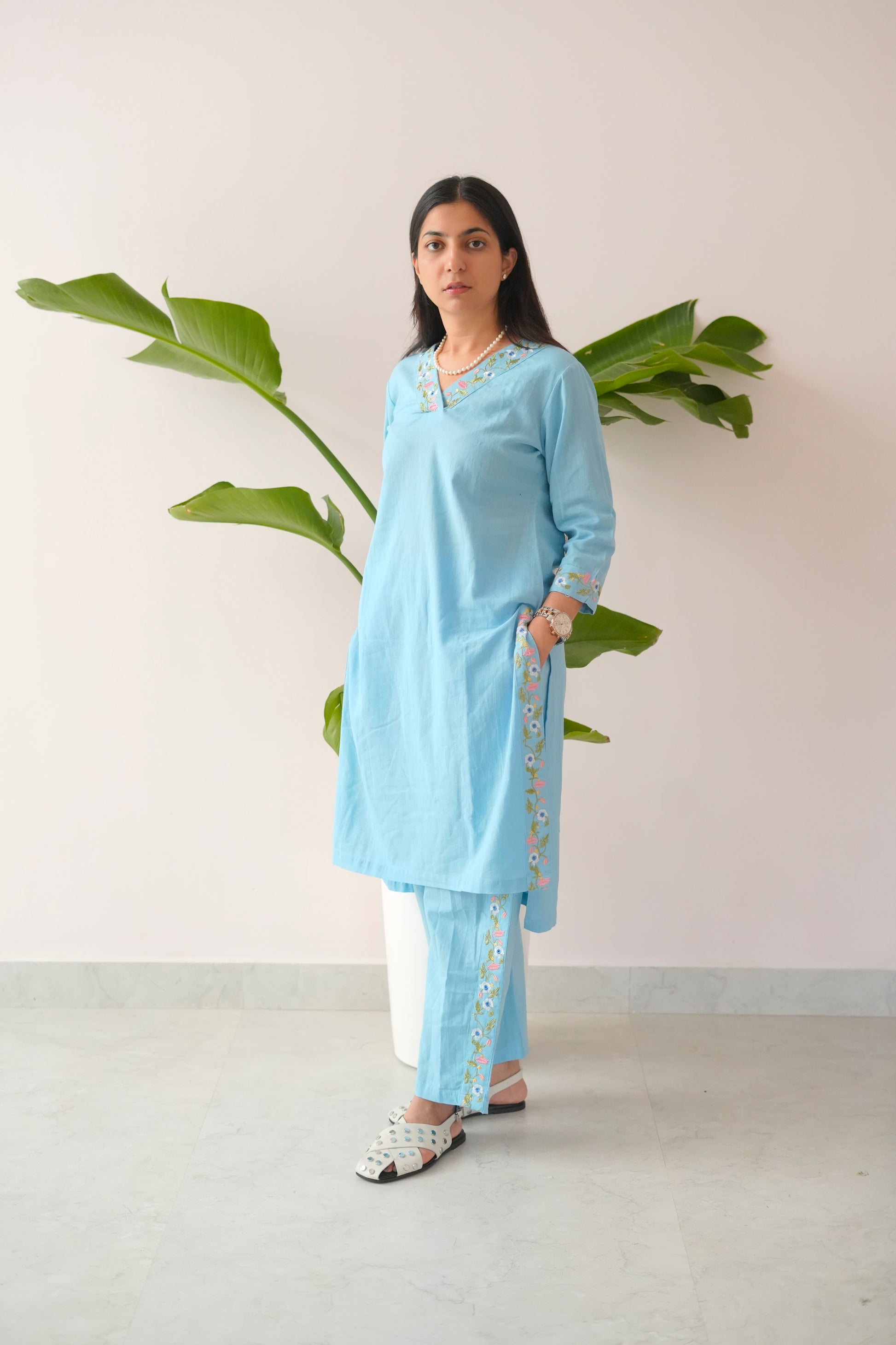 Woman wearing a light blue kurta with white pants standing against a plain background with a plant.