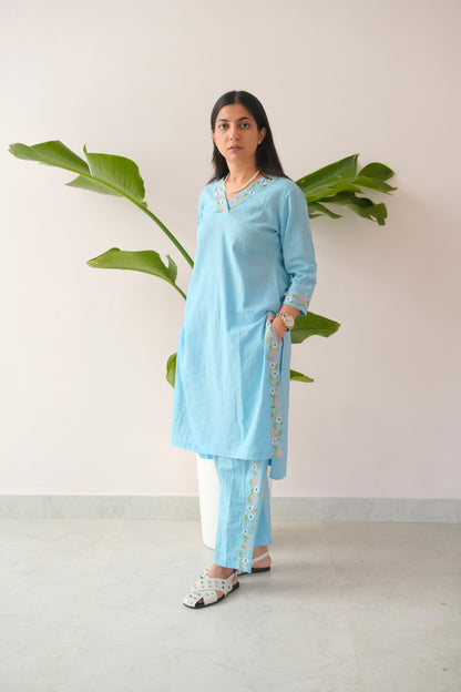 Woman wearing a light blue kurta with white pants standing against a plain background with a plant.
