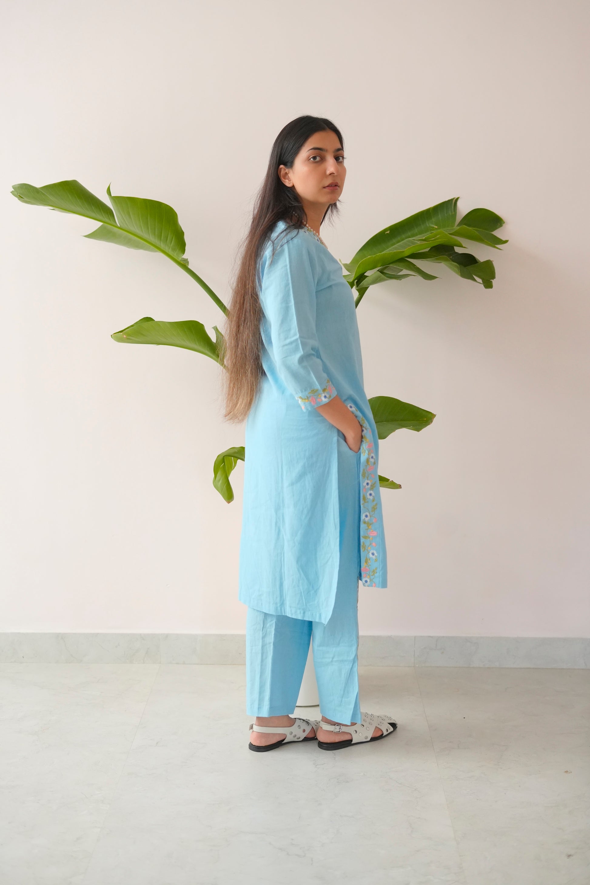 Woman wearing a light blue kurta with white borders standing next to a plant against a plain wall.