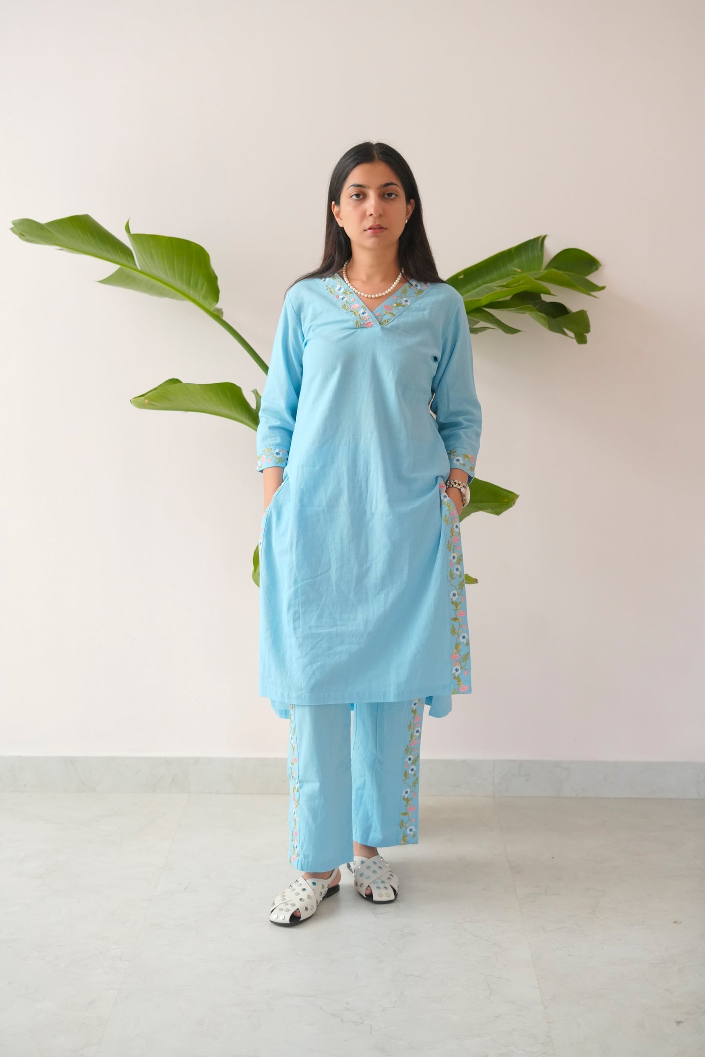 Woman wearing a light blue traditional outfit with white patterns, standing against a plain background with green leaves.