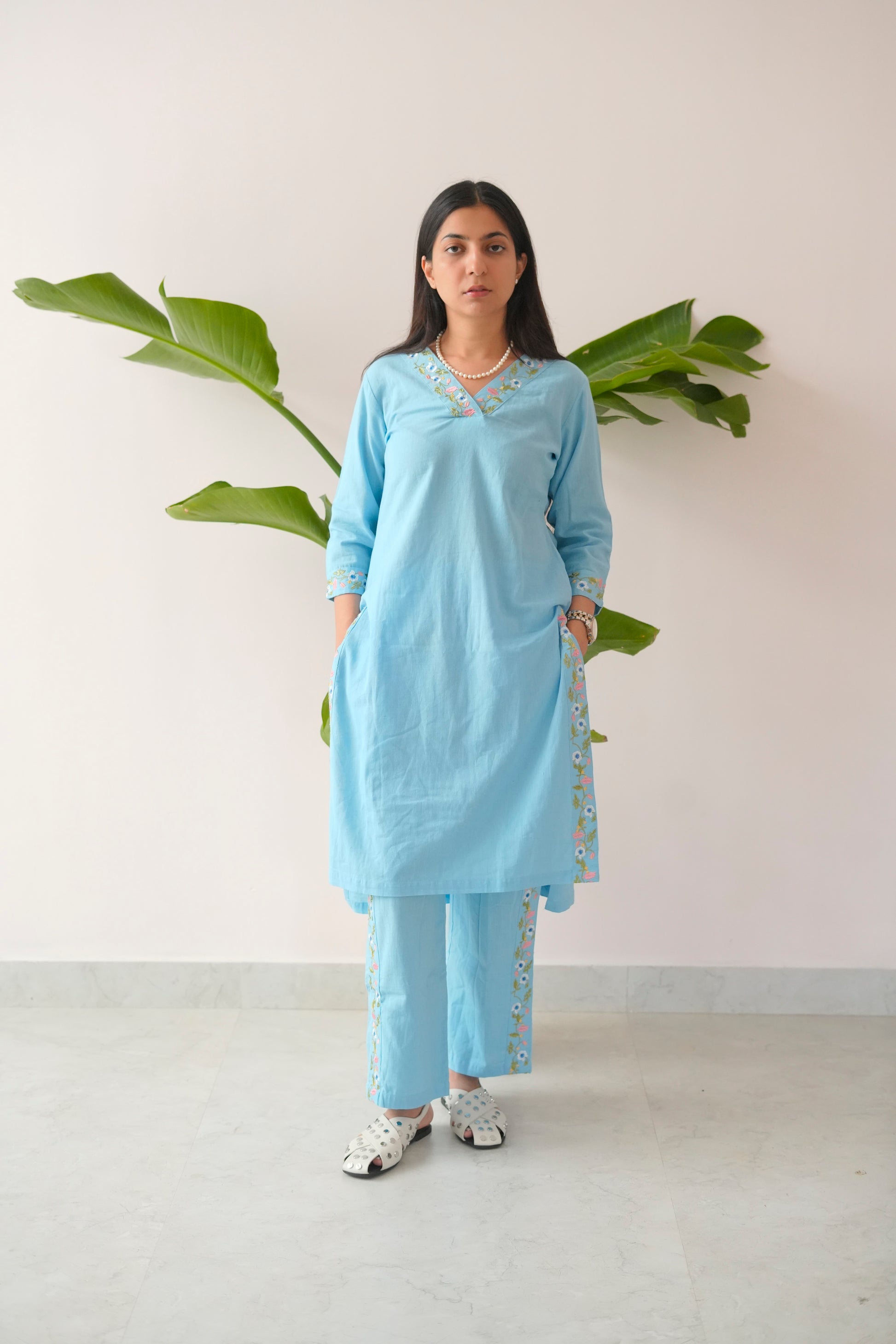 Woman wearing a light blue traditional outfit with white patterns, standing against a plain background with green leaves.