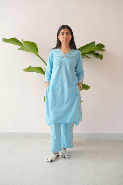 Woman wearing a light blue traditional outfit with white patterns, standing against a plain background with green leaves.