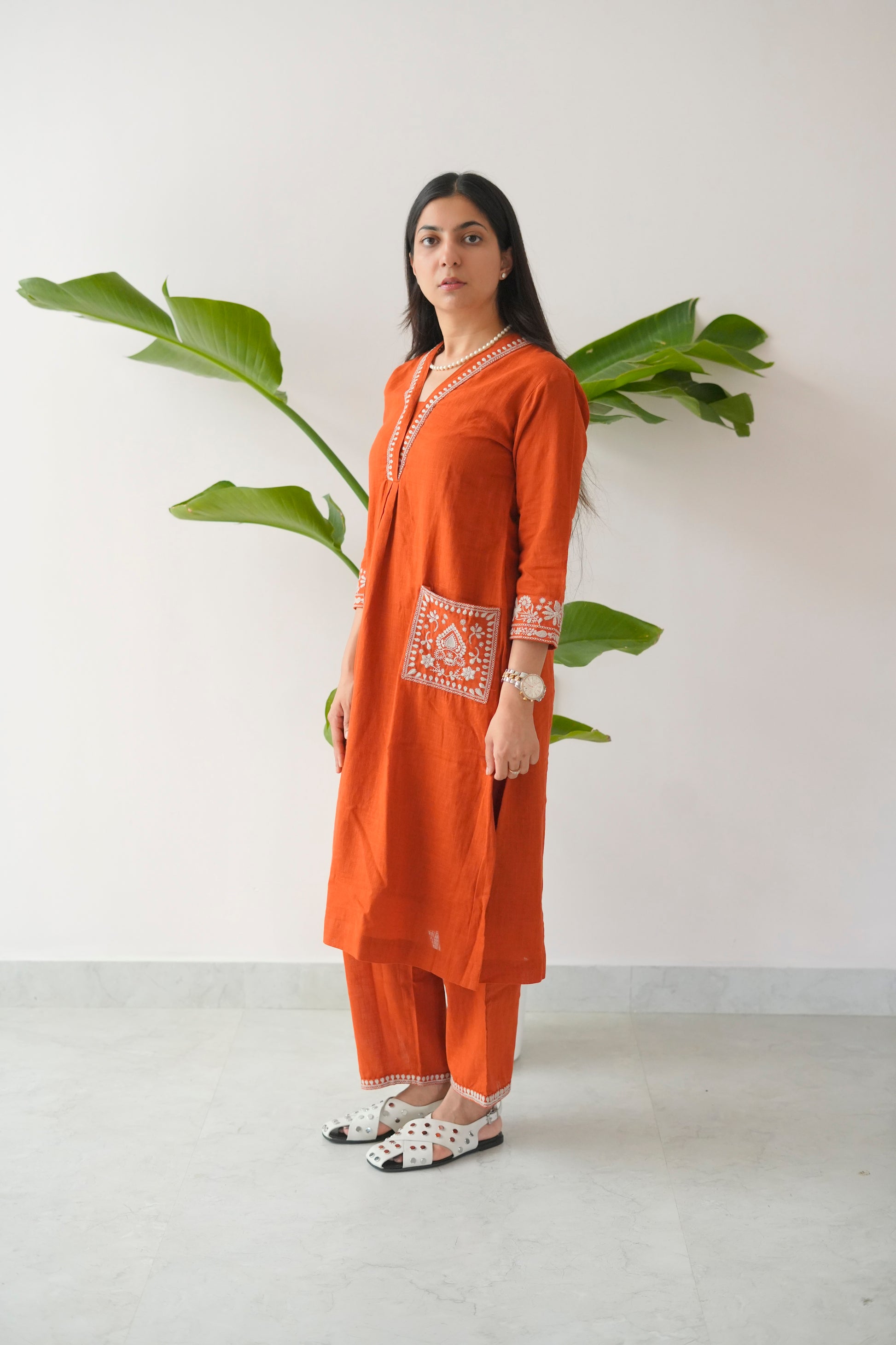 Woman wearing an orange traditional outfit with white patterns, standing against a plain background with a plant.