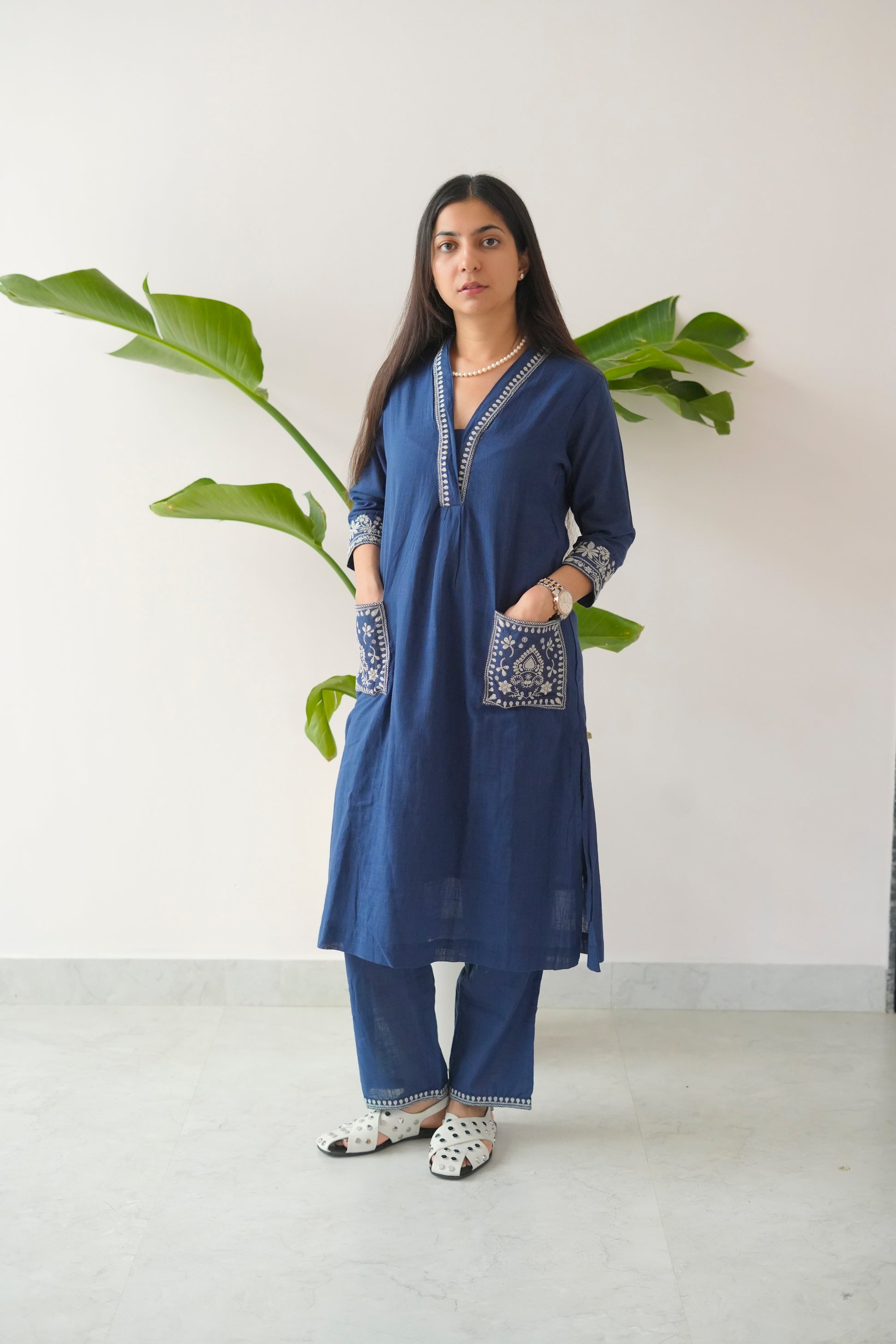 Woman wearing a blue embroidered kurta with white pants standing against a white wall with green leaves.
