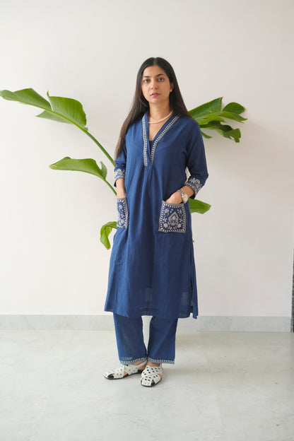 Woman wearing a blue embroidered kurta with white pants standing against a white wall with green leaves.