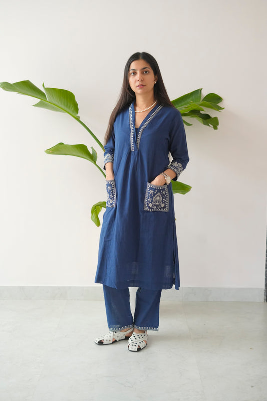 Woman wearing a blue embroidered kurta with white pants standing against a white wall with green leaves.