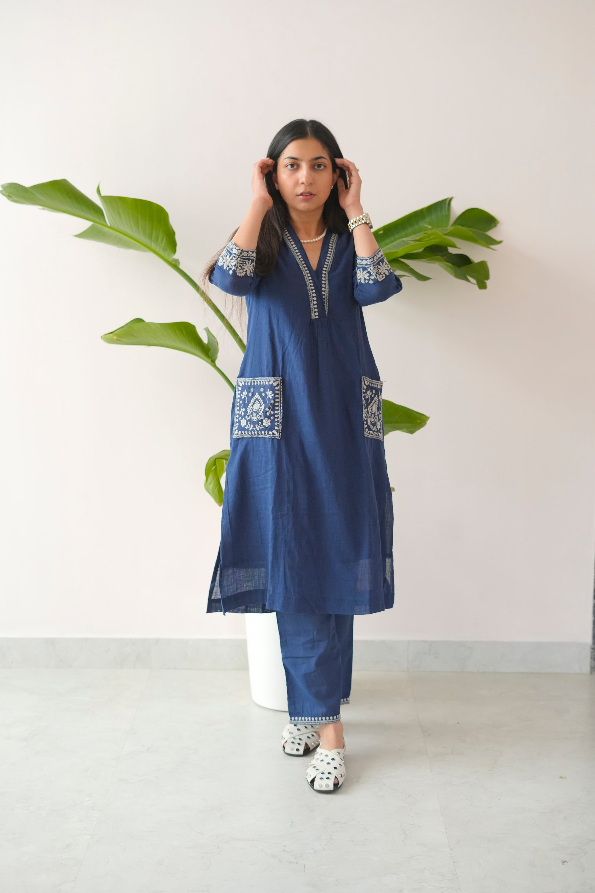 Woman wearing a blue embroidered kurta with white pants, standing against a white wall with green leaves.