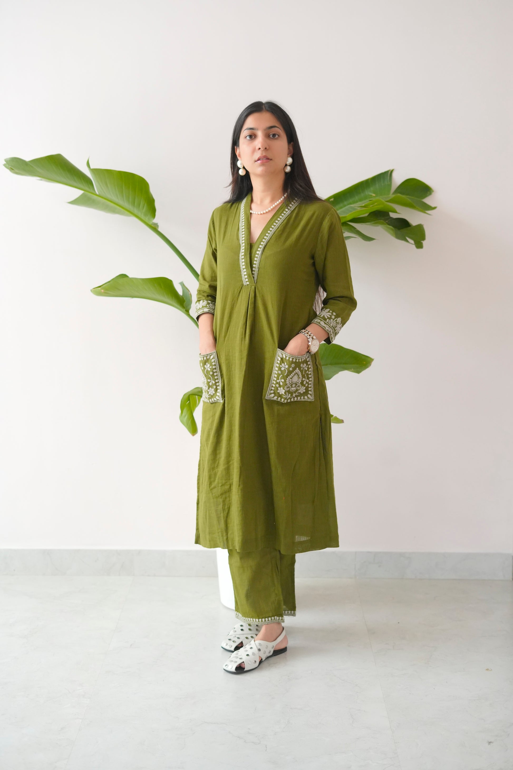 Woman wearing a green traditional outfit with white patterns, standing against a white wall with a plant.