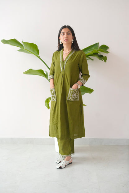 Woman wearing a green traditional outfit with white patterns, standing against a white wall with a plant.
