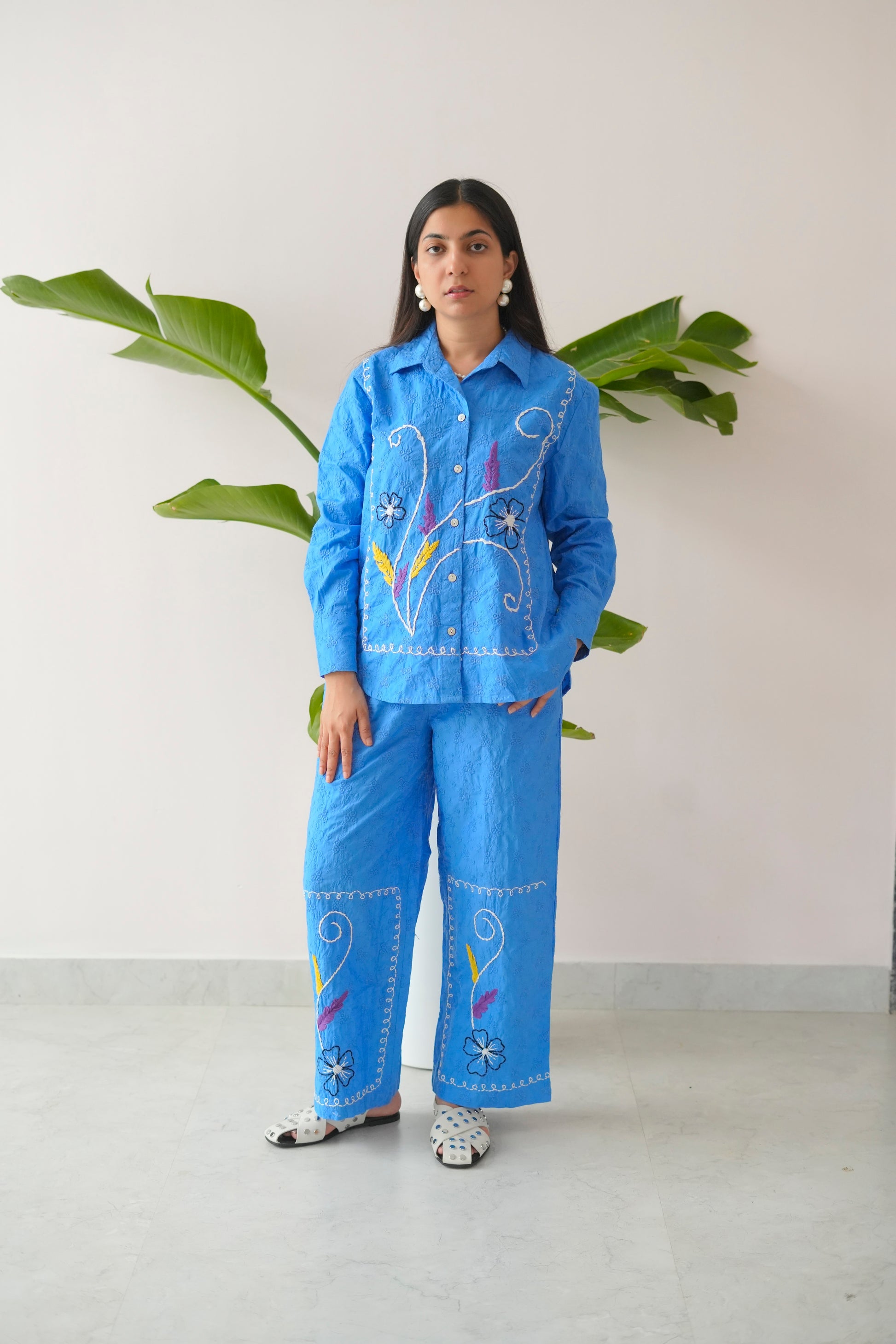 Person wearing a blue embroidered outfit standing against a white wall with green leaves.