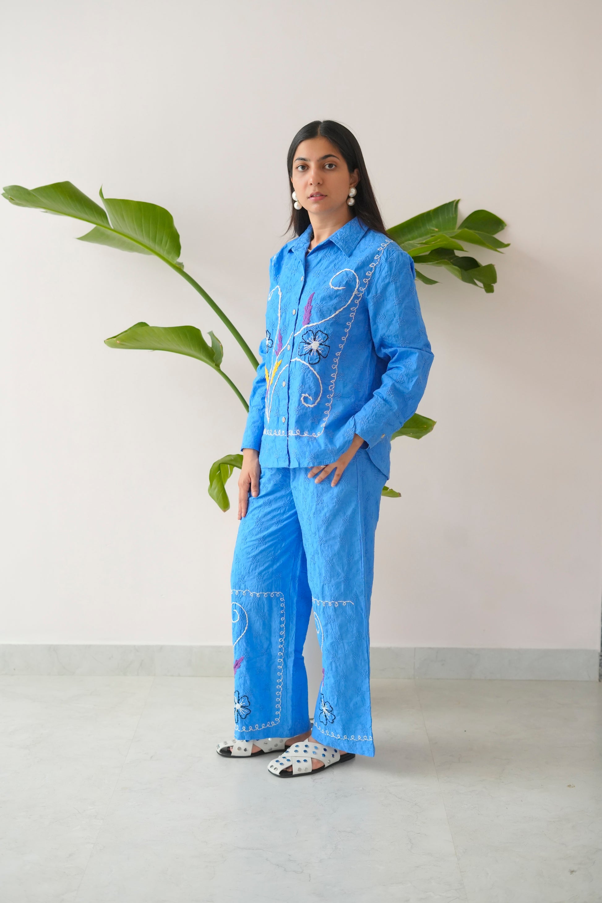 Woman wearing a blue embroidered outfit standing in front of a plant against a white wall.