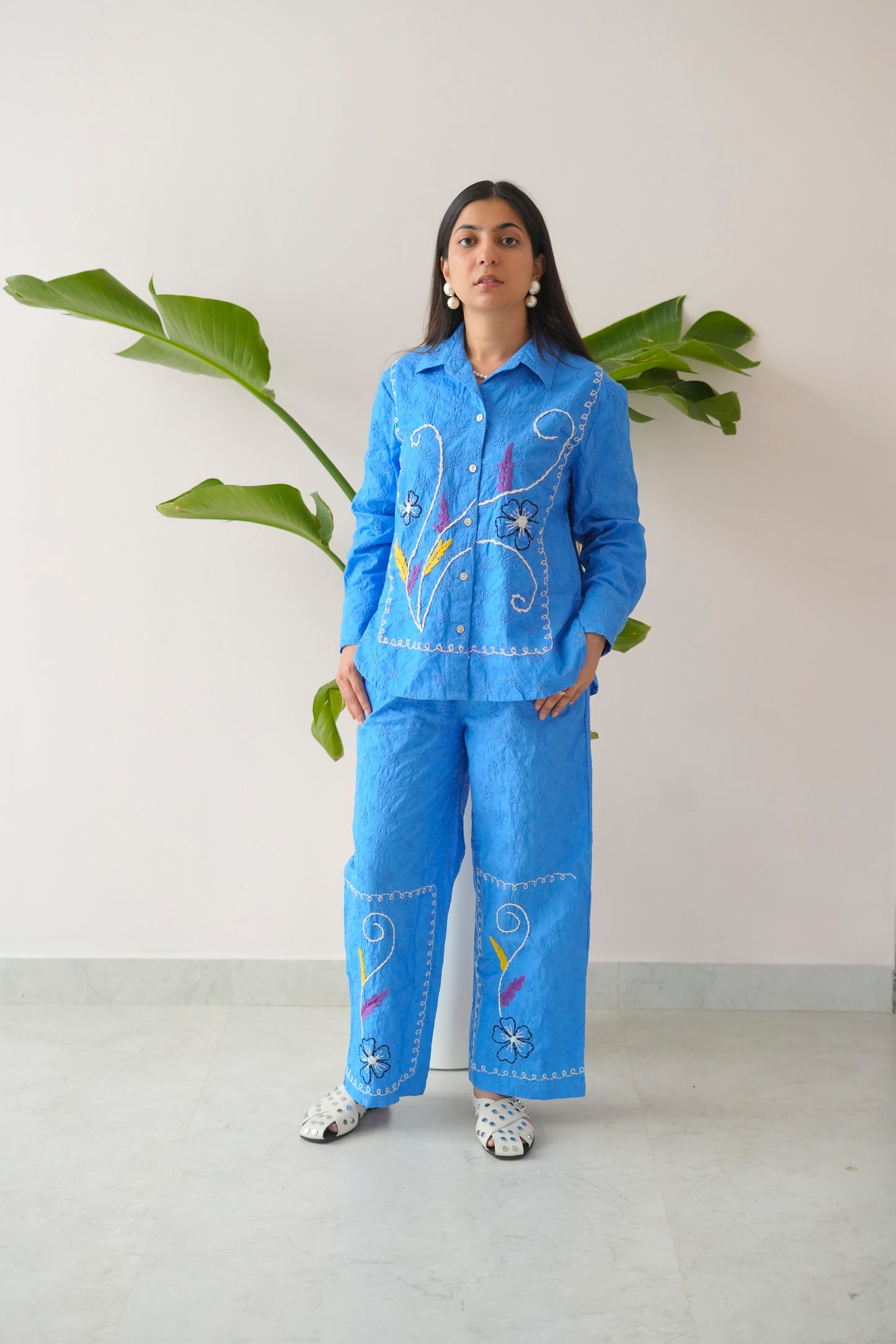 Person wearing a blue embroidered outfit standing against a white wall with a plant.
