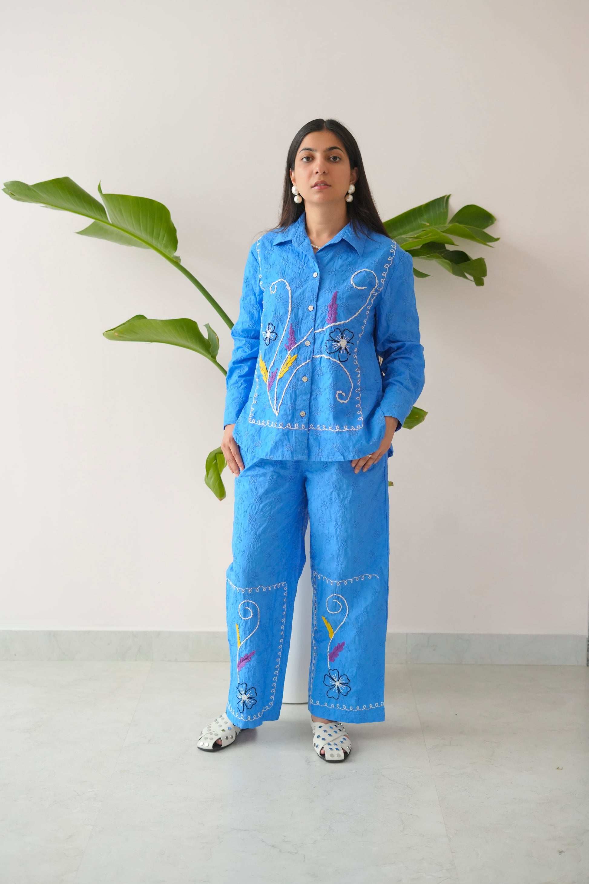 Person wearing a blue embroidered outfit standing against a white wall with a plant.