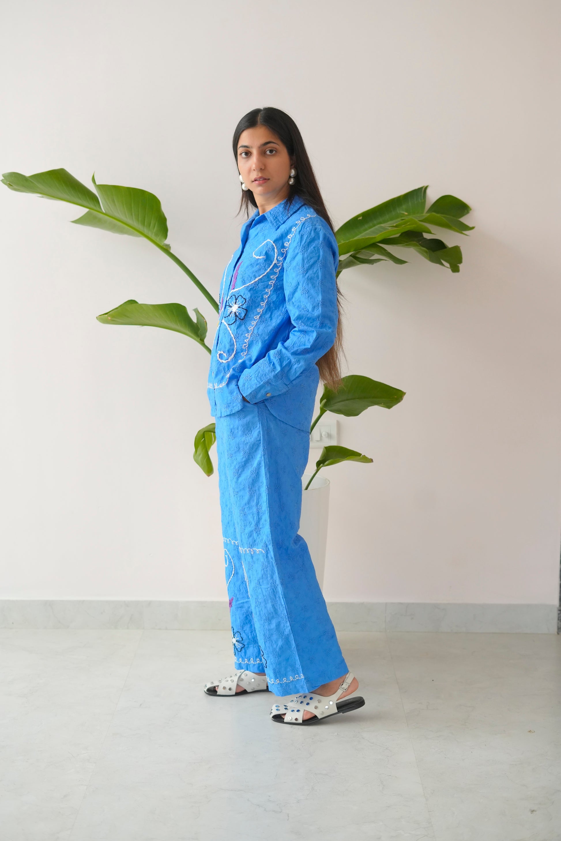 Person wearing a blue outfit with floral details standing next to a plant against a white wall.