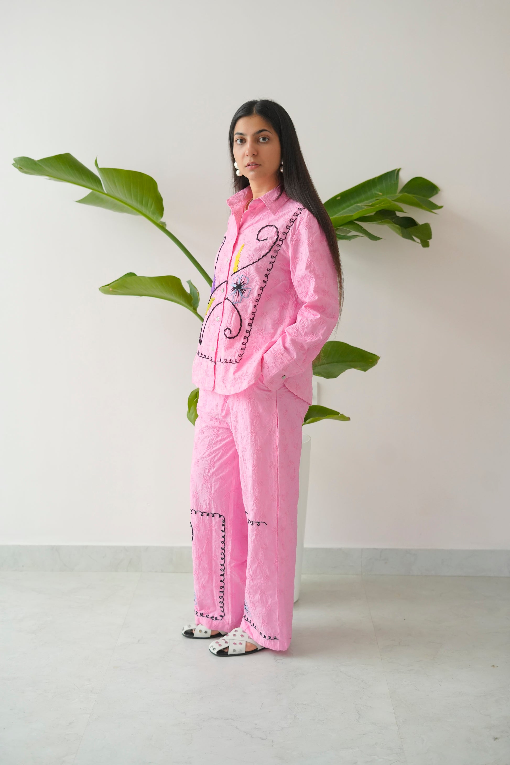 Person wearing a pink outfit with decorative elements, standing next to a plant against a white wall.