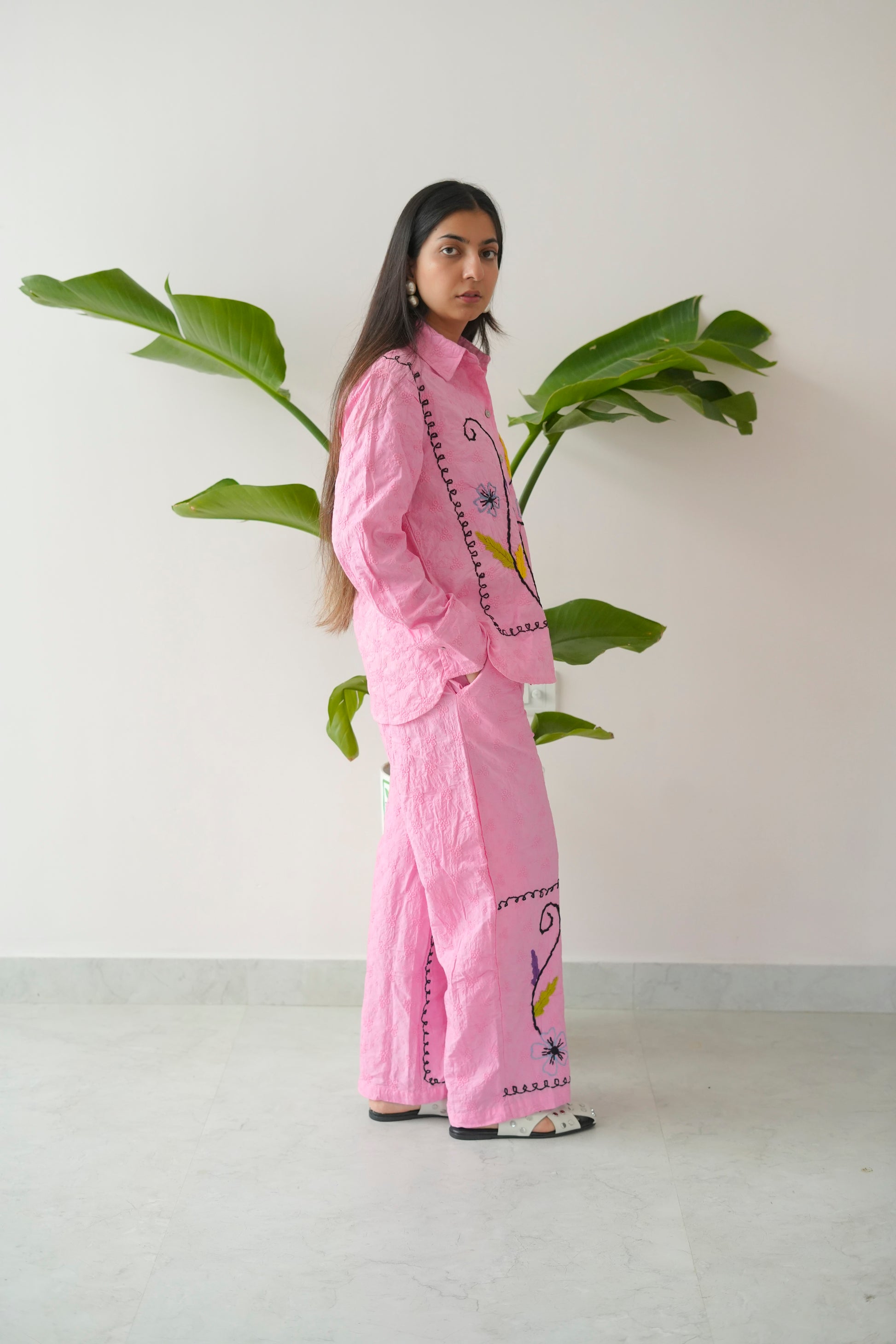 Person wearing a pink outfit with floral patterns standing next to a plant against a white wall.