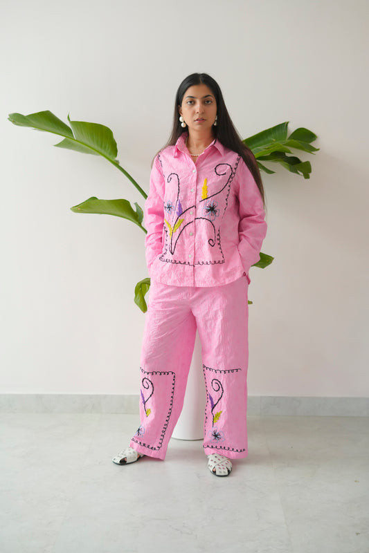 Person wearing a pink embroidered outfit standing against a white wall with green plants.