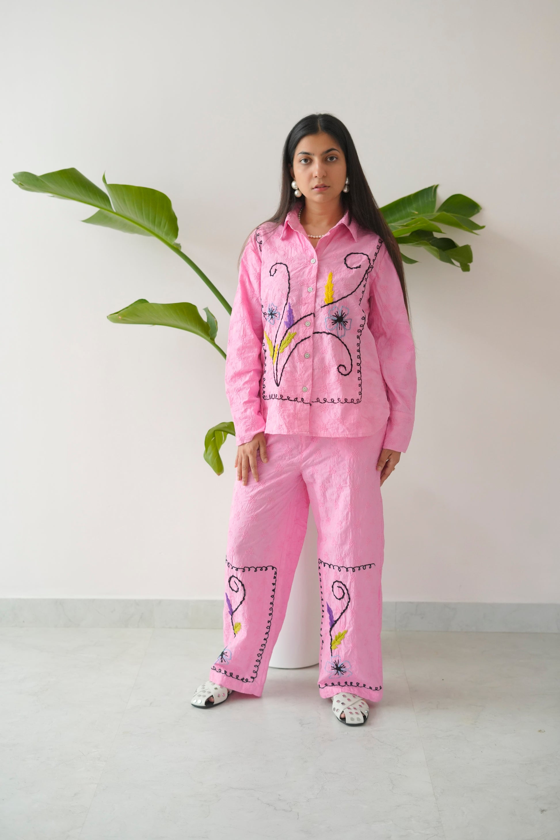 Person wearing a pink embroidered outfit standing against a white wall with green leaves.