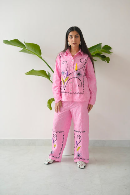 Person wearing a pink embroidered outfit standing against a white wall with green leaves.