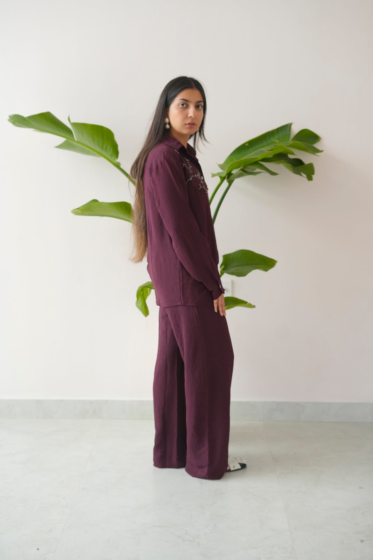 Person wearing a dark purple outfit standing next to a large green leafy plant against a white wall.
