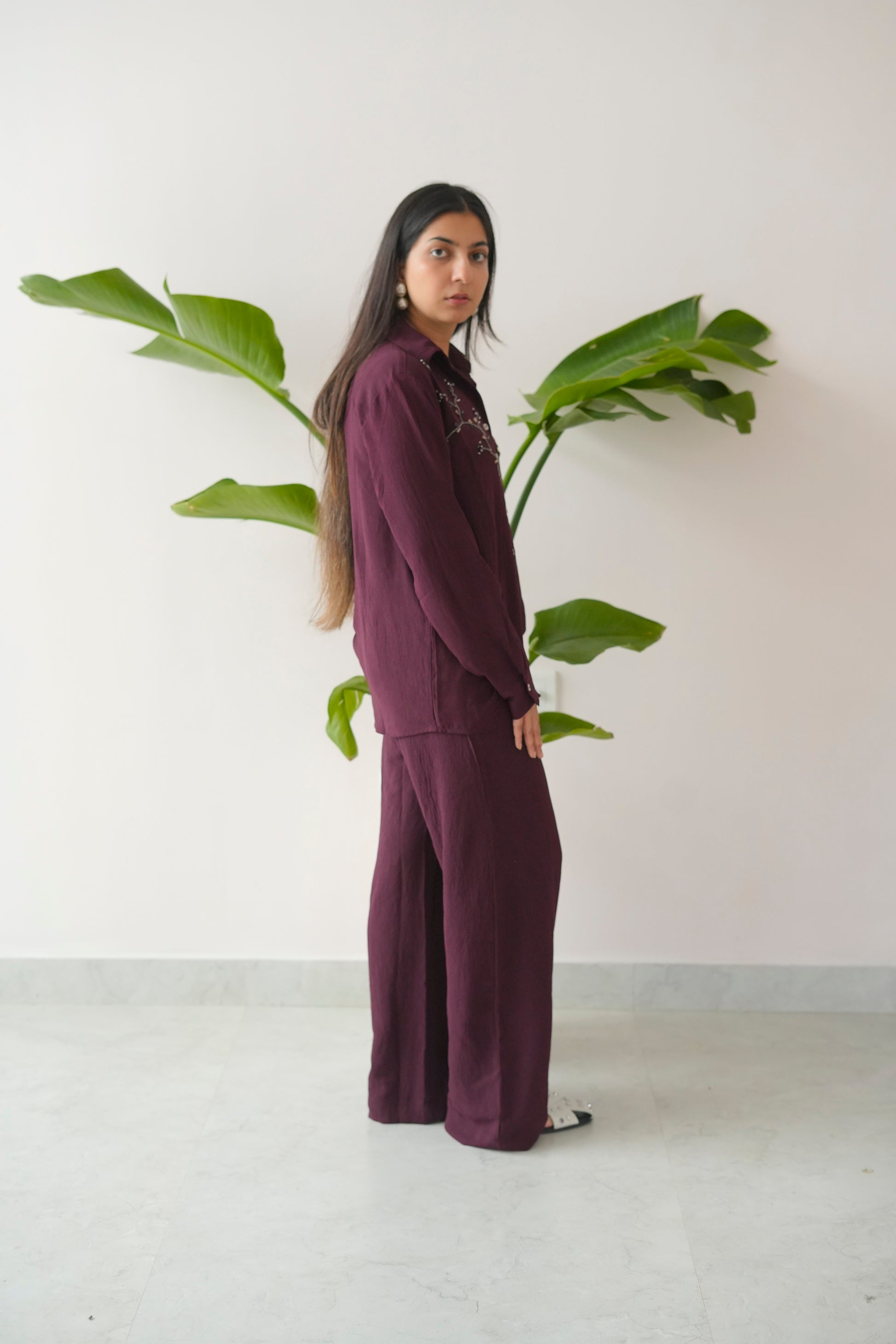 Person wearing a dark purple outfit standing next to a large green leafy plant against a white wall.
