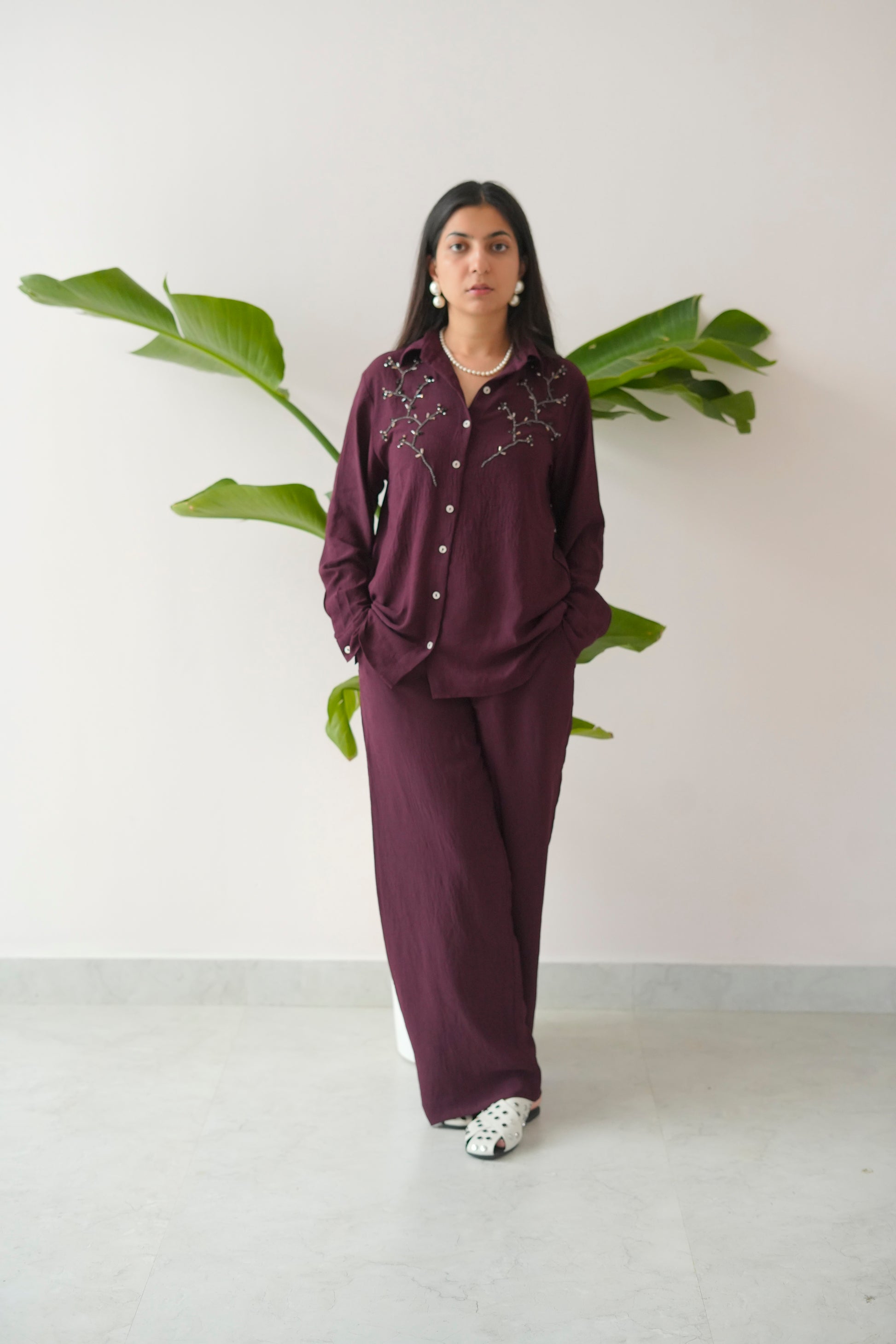 Woman wearing a dark purple outfit with white patterns, standing against a plain background with a plant.