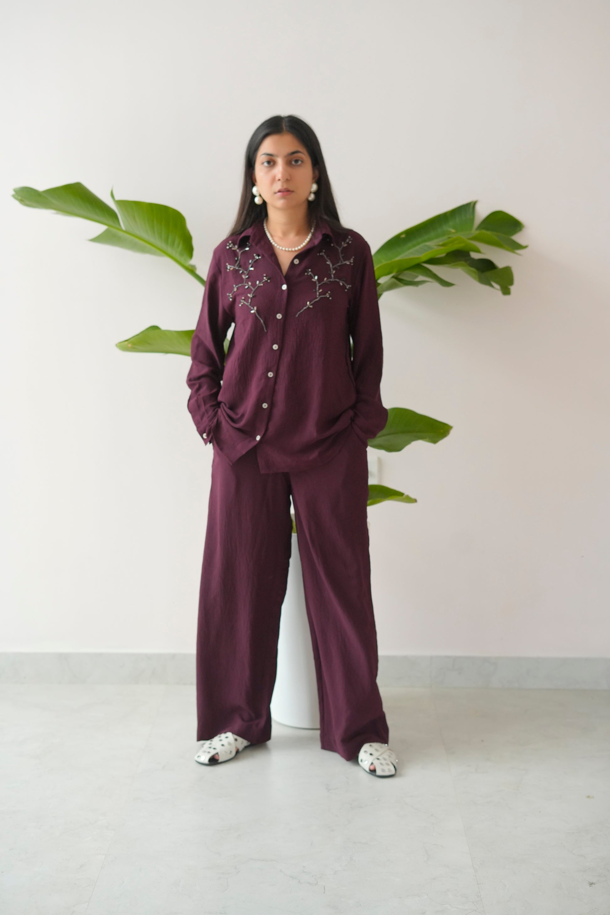 Person wearing a burgundy outfit with floral embroidery in a minimalistic setting with plants.