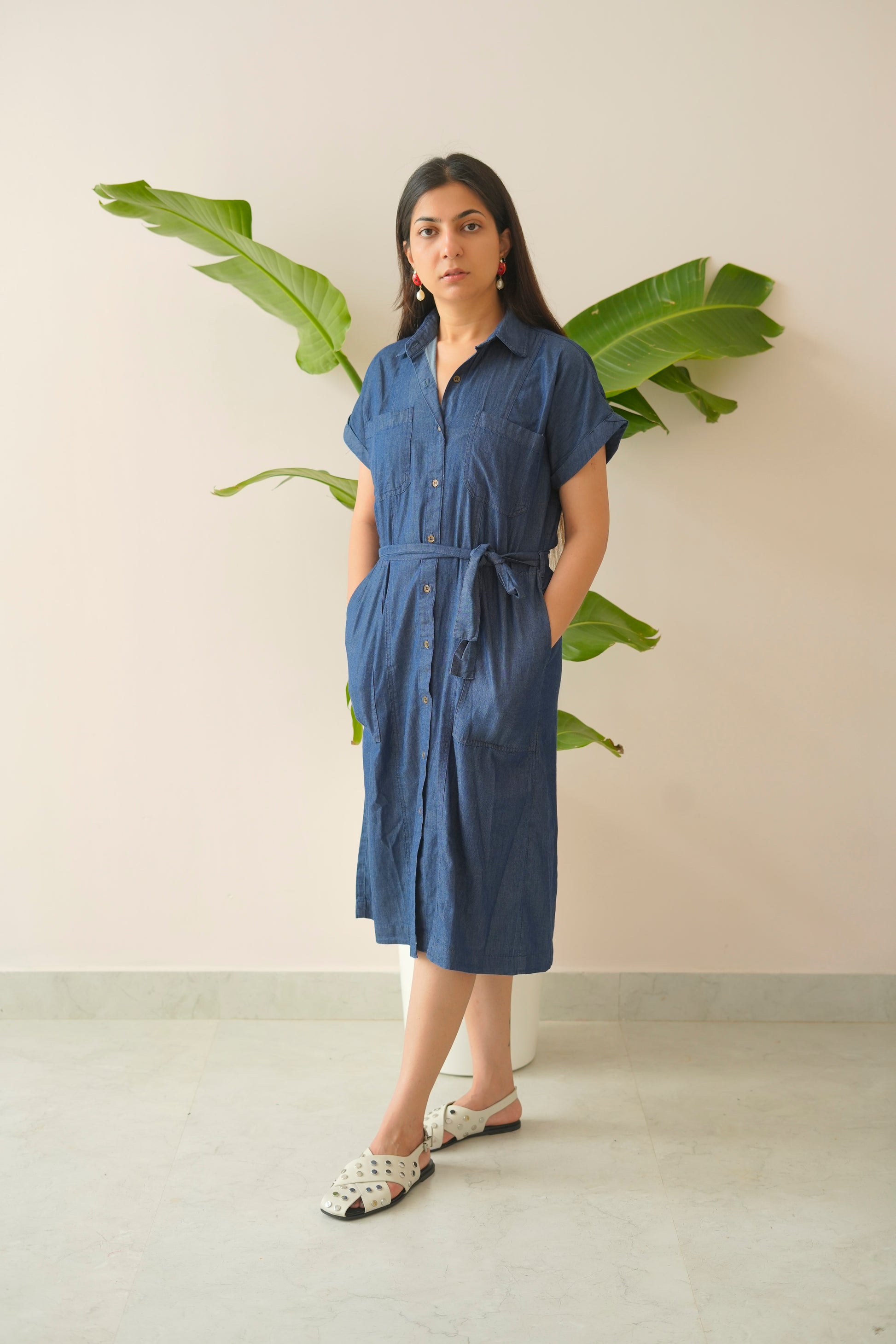 Woman wearing a blue dress standing against a light background with green leaves.