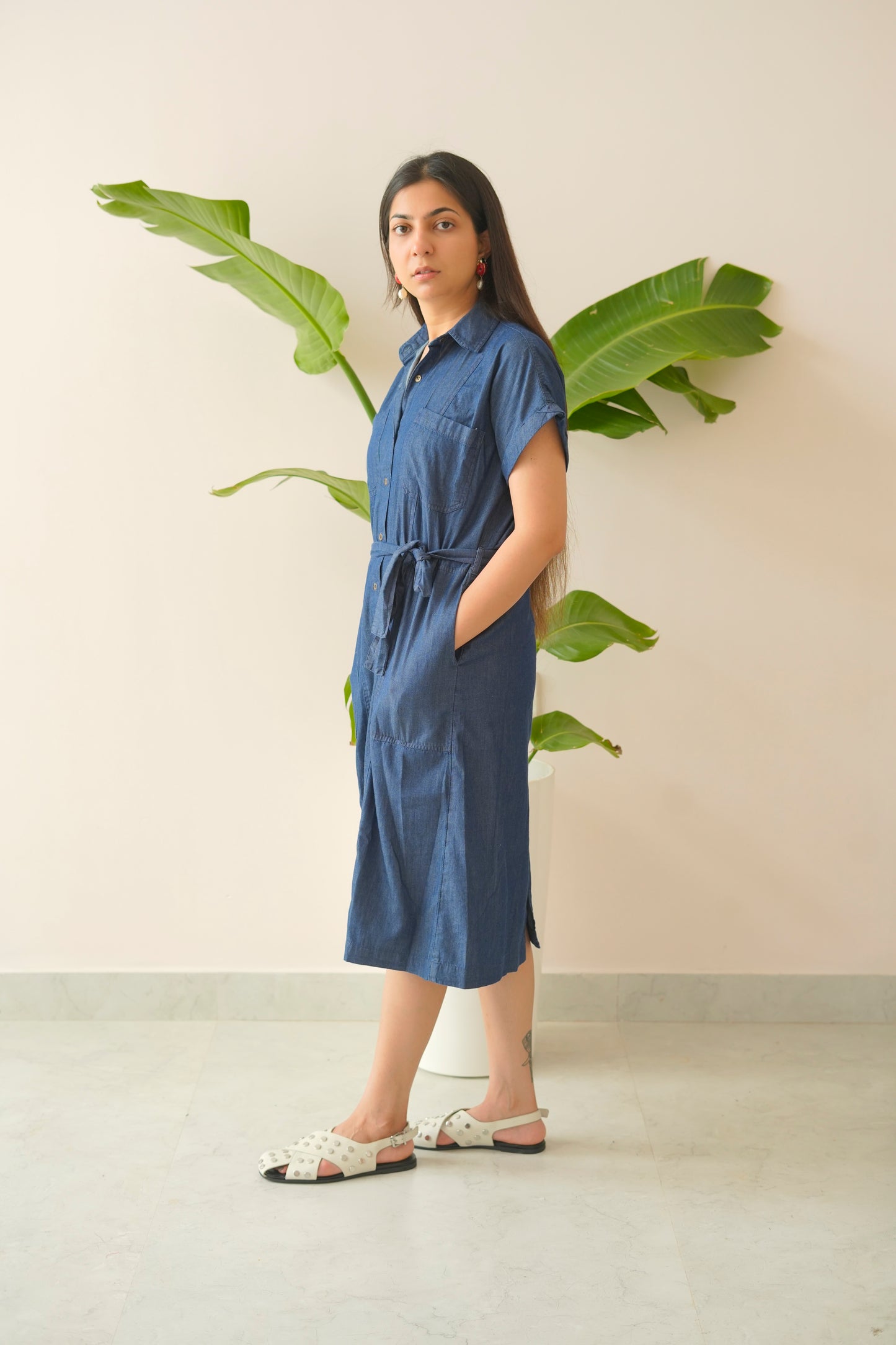 Woman wearing a blue dress standing in front of a plant against a beige wall.
