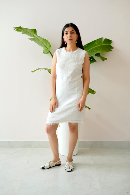 Woman wearing a white dress standing against a plain background with a plant.