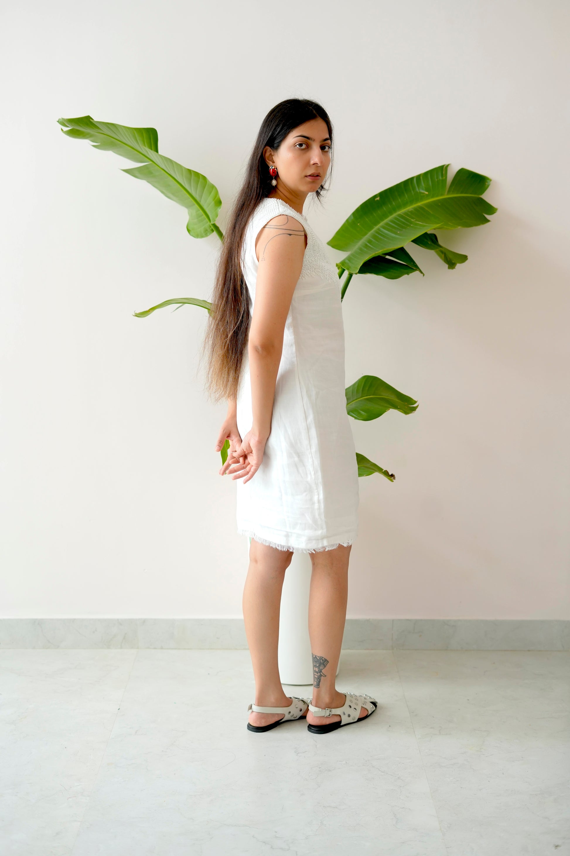 Woman in a white dress standing between two large green leaves against a plain wall.