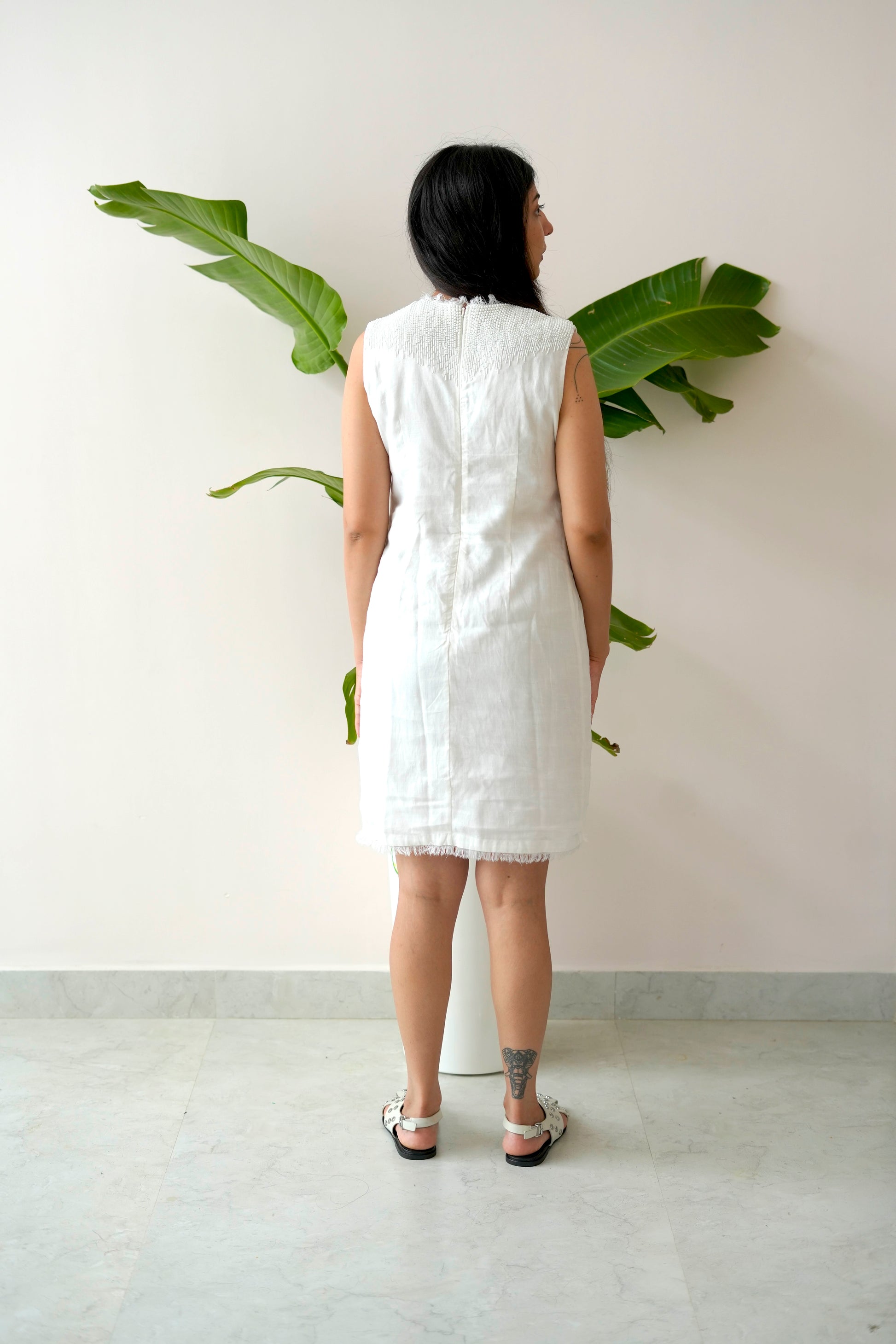 Person wearing a white sleeveless dress standing against a plain wall with green plants.