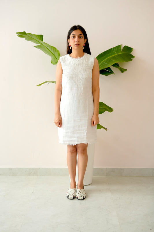 Woman wearing a white dress standing against a light pink wall with a plant.